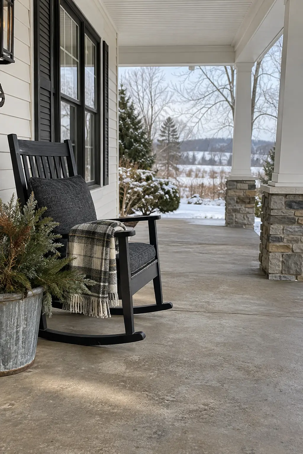 Covered porch with one black rocking chair, plaid throw, and evergreen bucket